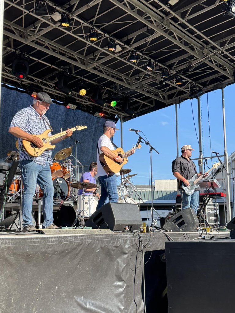 A four-piece band performs on a large outdoor stage at the Red Rose Festival
