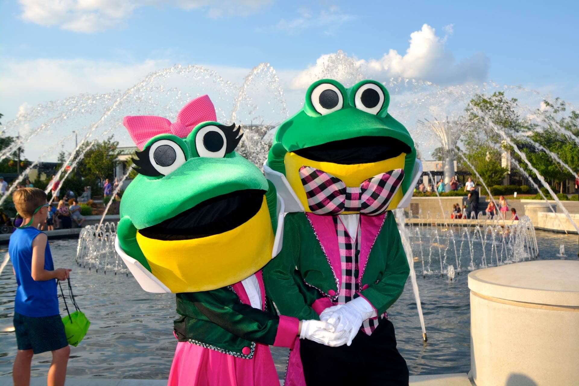 Two people in colorful frog mascot costumes pose together in front of a fountain during the Come-See-Me Festival, with families enjoying the outdoor event in the background.