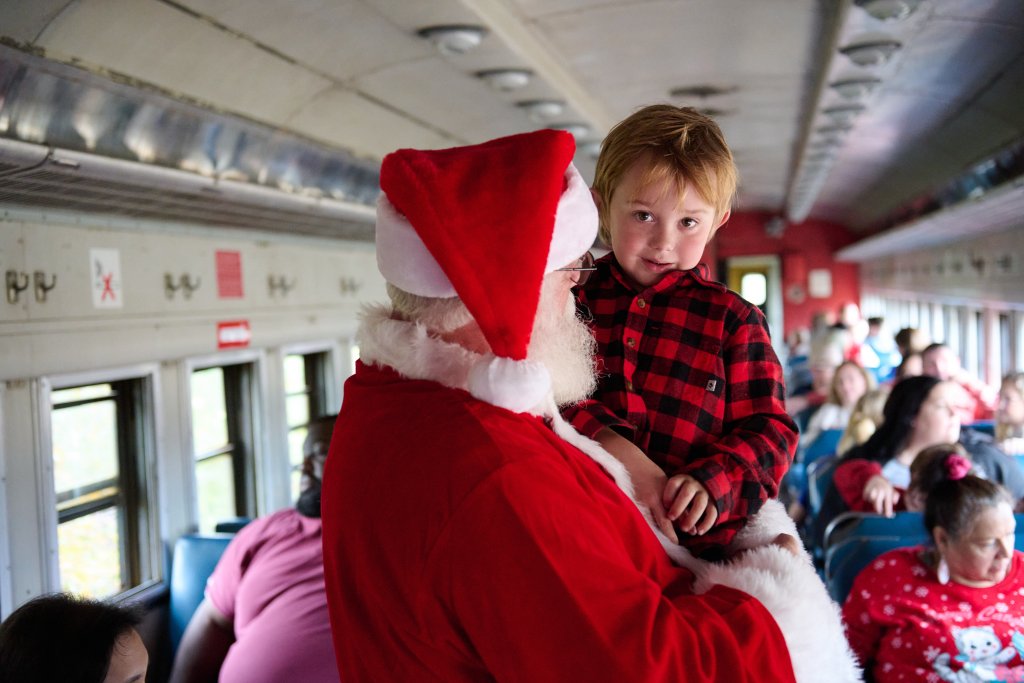 Santa Claus holds a young child dressed in a red plaid shirt aboard a festive train ride at the South Carolina Railroad Museum, surrounded by families seated in the vintage railcar.