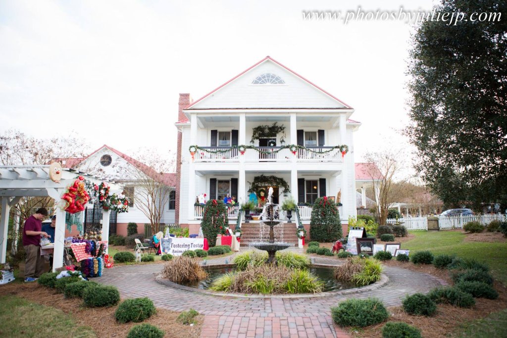 A large white Southern-style home decorated with garlands, wreaths, and Christmas displays hosts an outdoor Country Christmas Shopping Trunk Show, with vendors and holiday décor arranged around a brick walkway and fountain in the front yard.