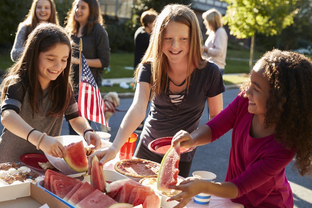 three girls smile at one another as they all grab a slice of watermelon during an event in the Olde English District