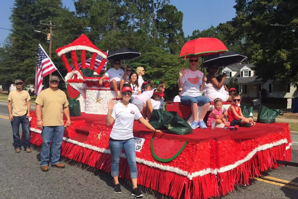 a parade float full of smiling and waving people is seen at a festival in the Olde English District