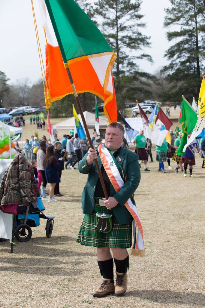 A man dressed in traditional Irish attire carries a large Irish flag during Irish Fest Camden, surrounded by festival goers and colorful banners on an open field.